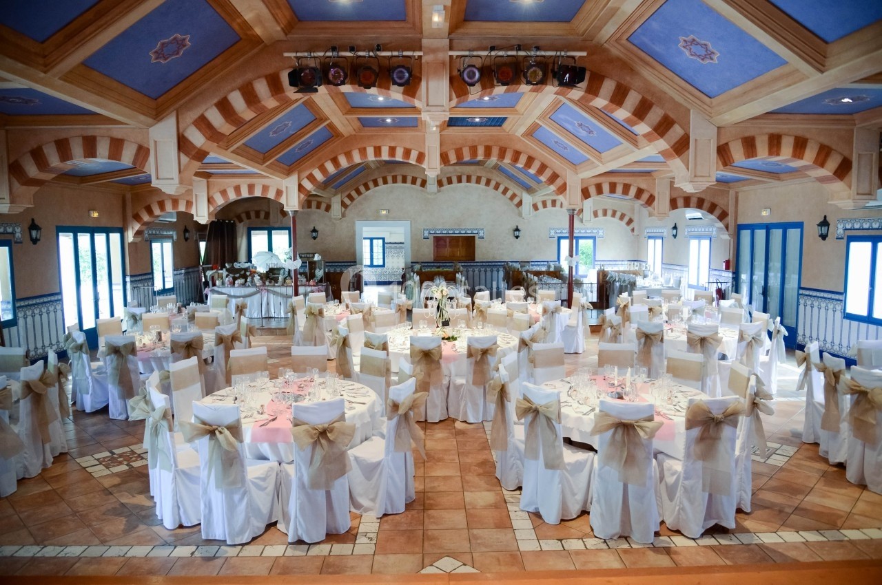 Salle de réception décorée pour un événement, avec des tables rondes dressées et des chaises ornées de nœuds.