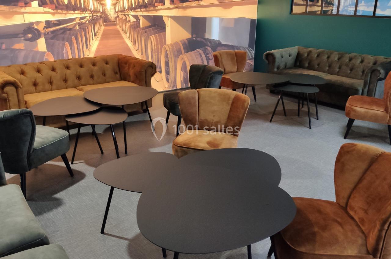 Salon avec fauteuils et canapés en velours, tables basses noires et décor mural représentant des fûts de bois empilés.