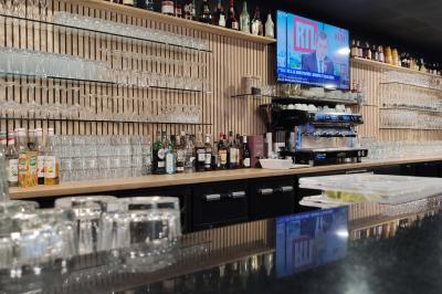 Bar moderne avec comptoir en marbre noir, étagères remplies de verres et bouteilles, machine à café et écran allumé.