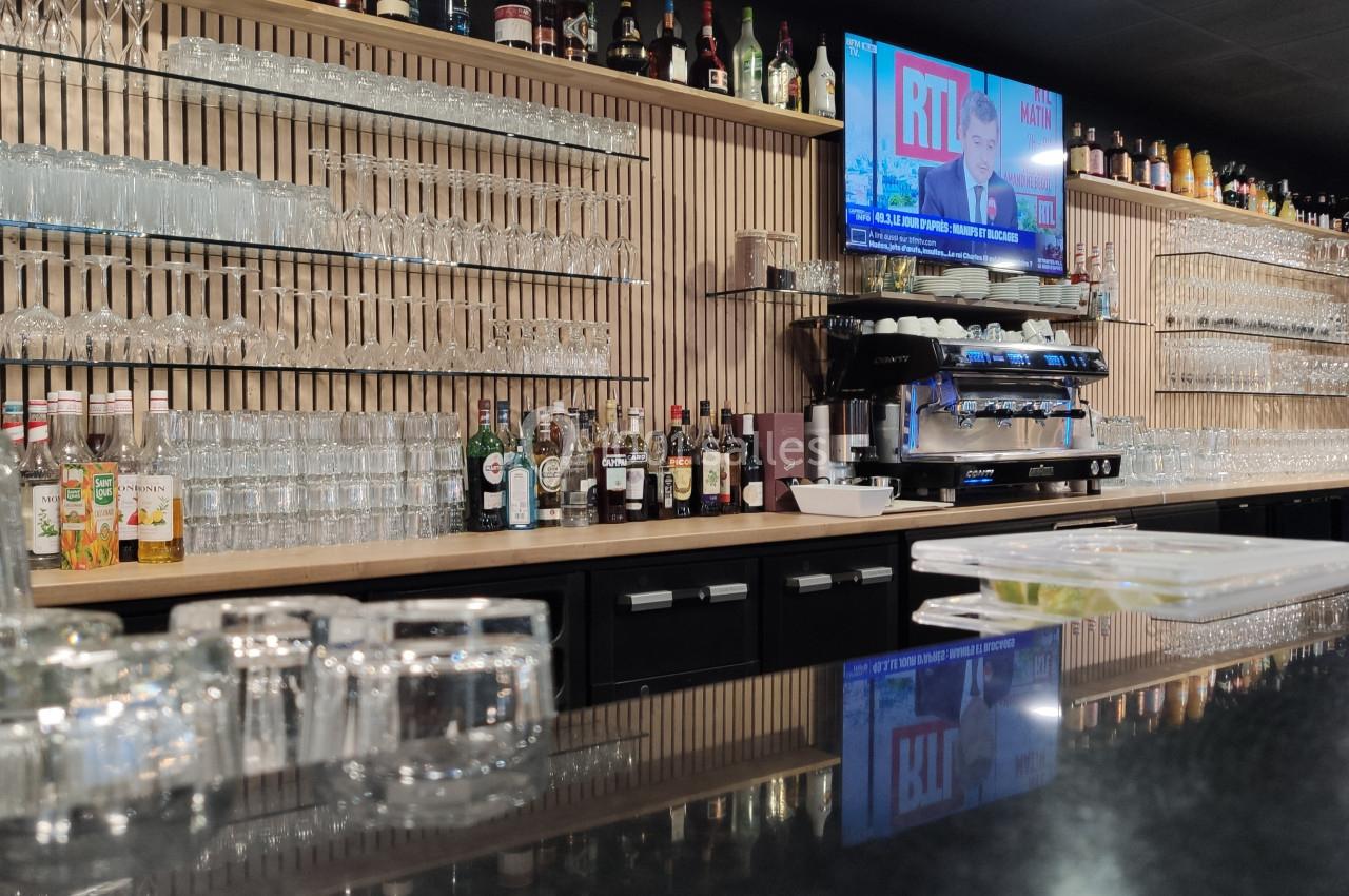 Bar moderne avec comptoir en marbre noir, étagères remplies de verres et bouteilles, machine à café et écran allumé.