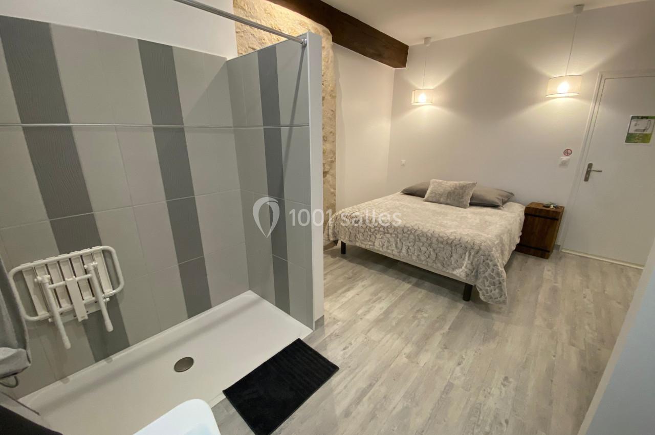Chambre avec lit double, douche accessible, sol en parquet clair et mur en pierre partiellement visible.