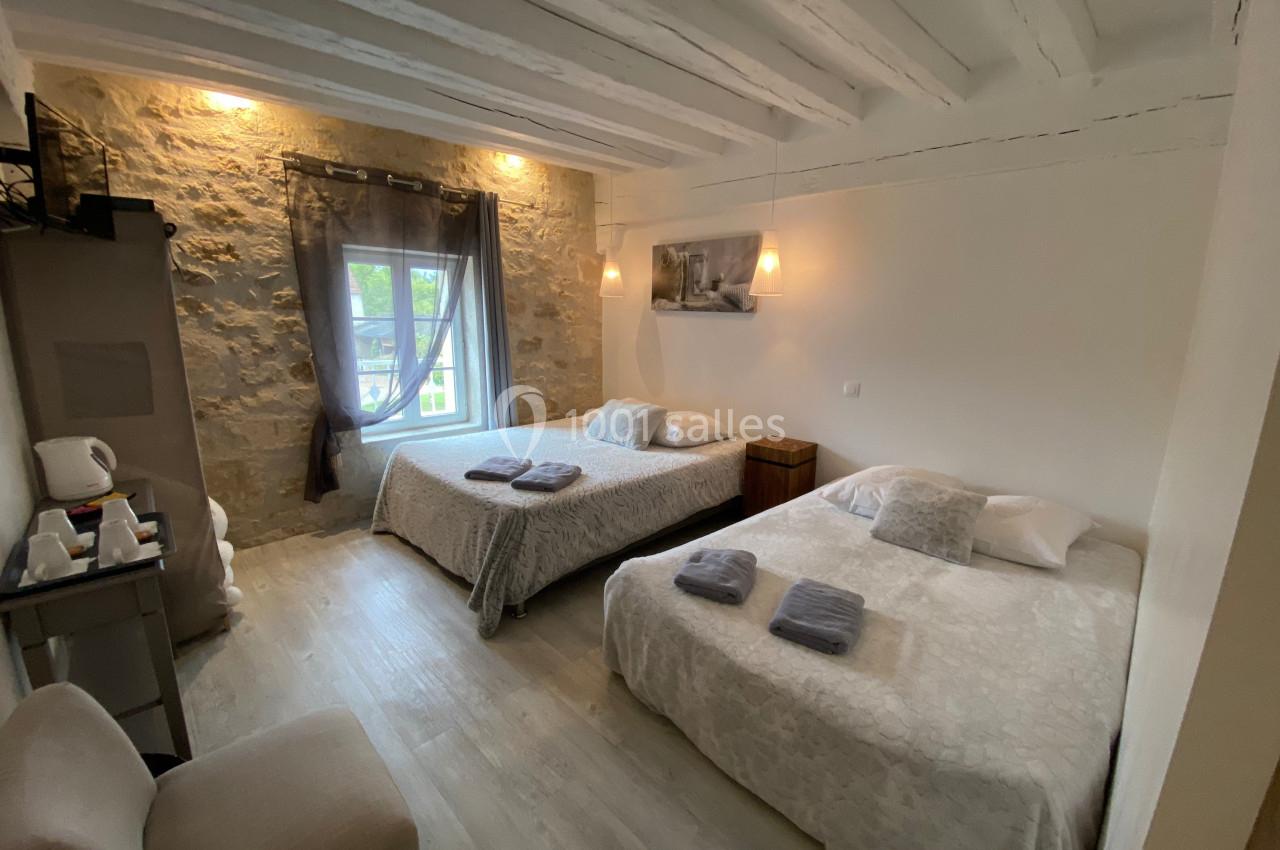 Chambre lumineuse avec deux lits doubles, murs en pierre apparente, fenêtre avec rideaux et mobilier simple.