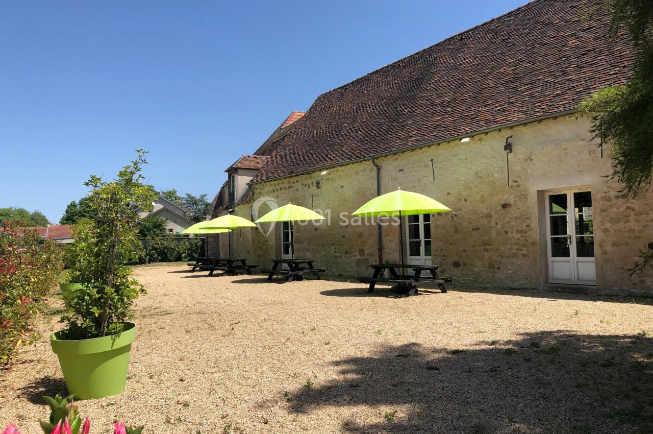 Cour extérieure en gravier avec tables de pique-nique et parasols verts, bordée d'un bâtiment en pierre sous un ciel bleu.