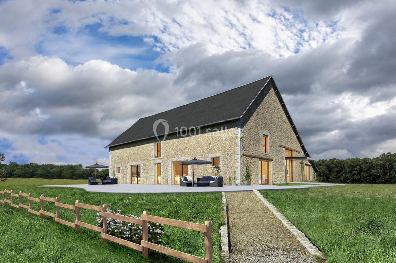 Grande maison en pierre avec toit en ardoise, entourée de verdure, avec terrasse aménagée et ciel nuageux en arrière-plan.