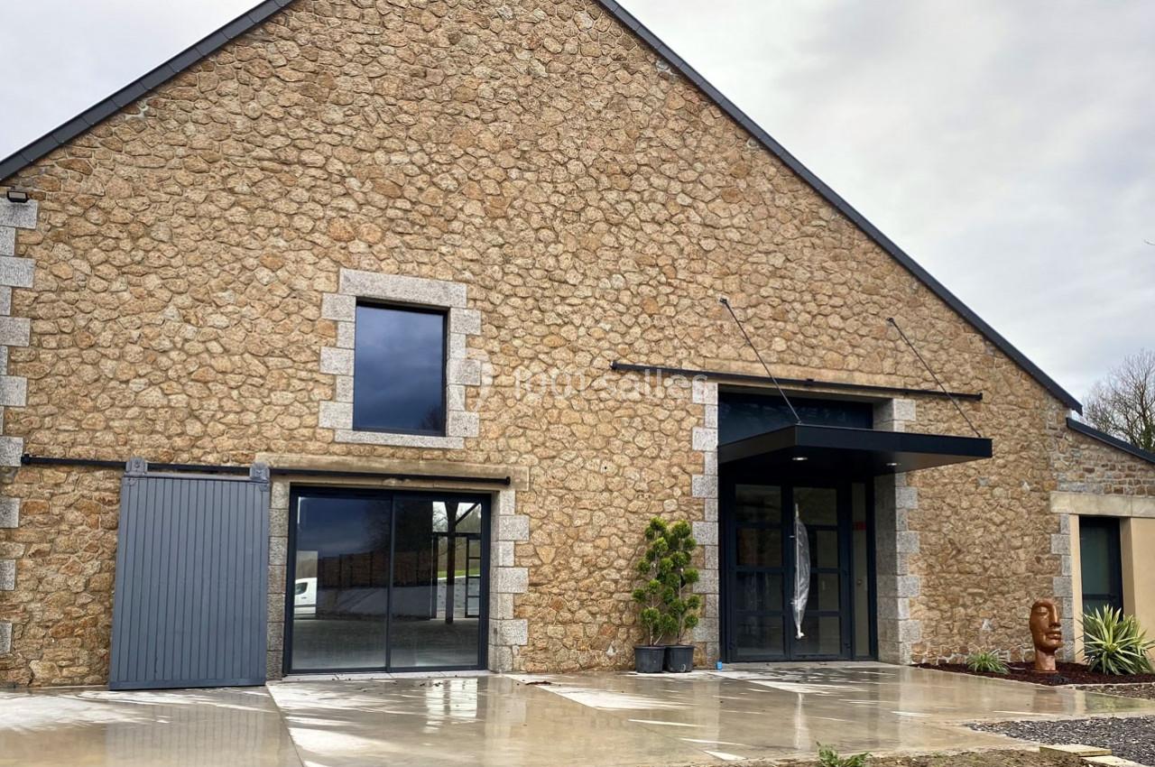 Façade en pierre d'un bâtiment avec grandes baies vitrées, porte d'entrée moderne et sol en béton mouillé.