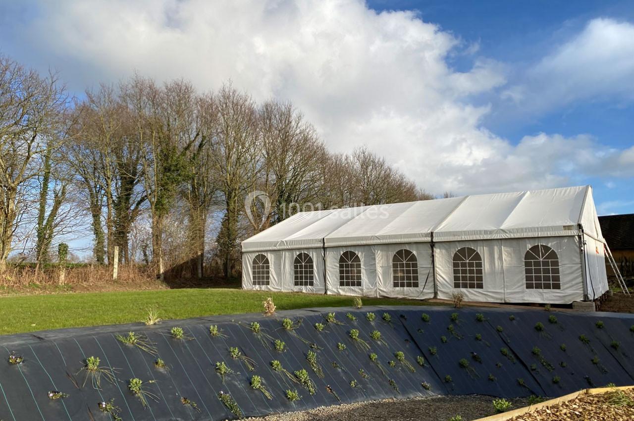 Grande tente blanche installée sur une pelouse près d'arbres, avec un talus végétalisé au premier plan.