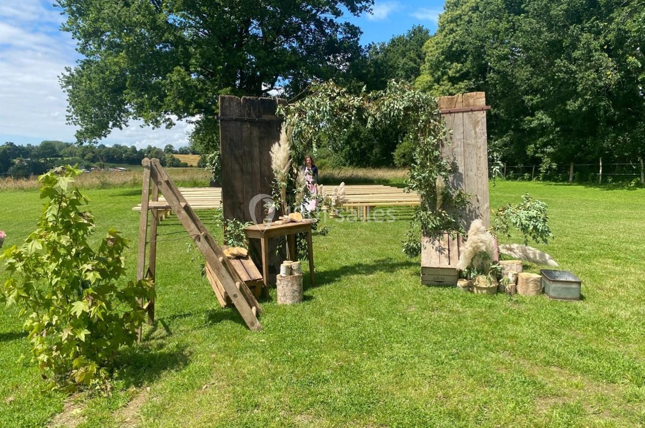Décor champêtre avec arche en bois ornée de feuillage, bancs en arrière-plan sur une pelouse entourée d'arbres.