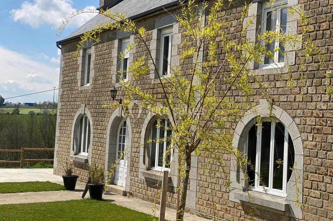 Façade en pierre d'une maison avec fenêtres cintrées, arbre jeune au premier plan et paysage rural en arrière-plan.