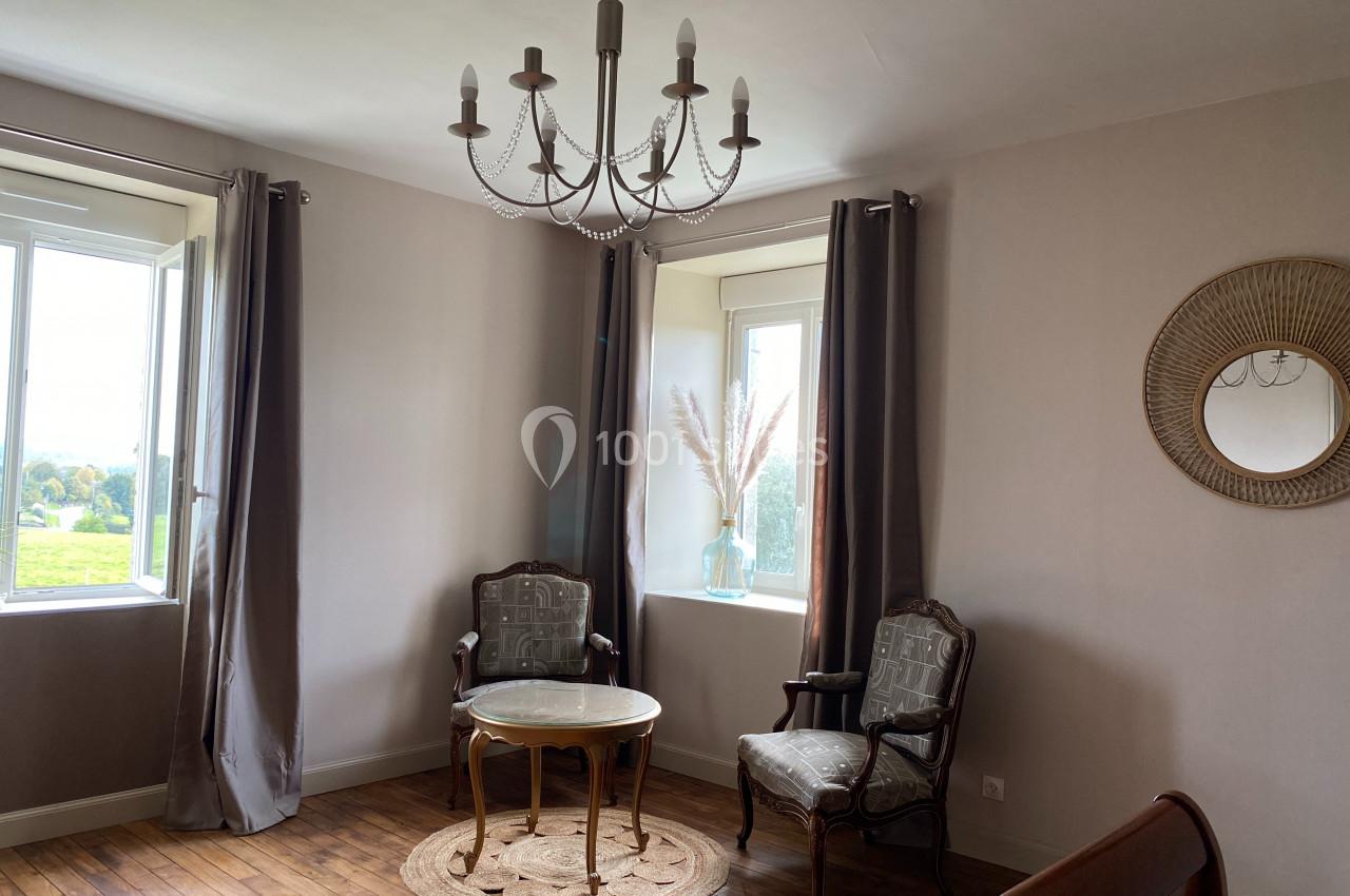 Deux fauteuils et une table ronde sur un tapis, près de deux fenêtres avec rideaux, dans une pièce lumineuse.