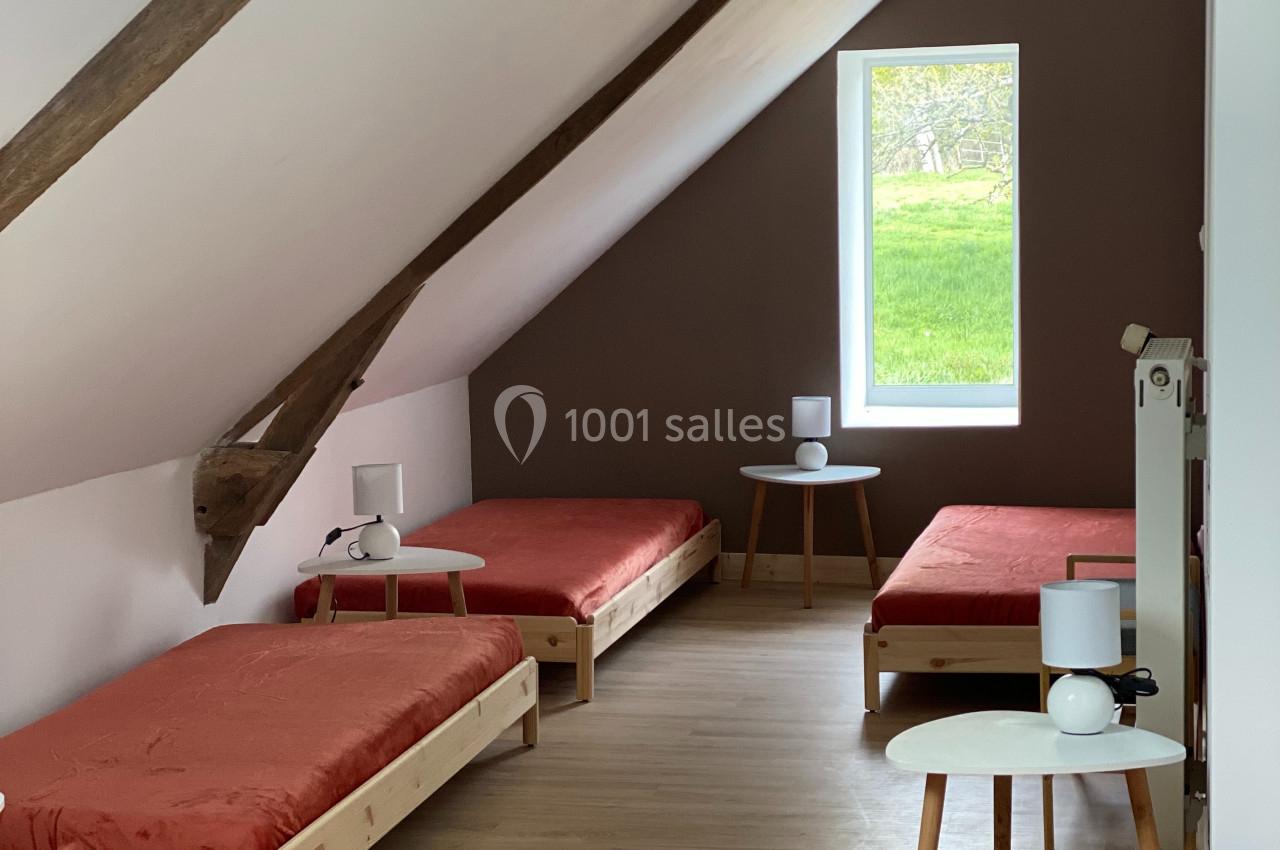 Quatre lits simples avec draps rouges dans une chambre mansardée lumineuse, avec murs blancs et une fenêtre donnant sur un…