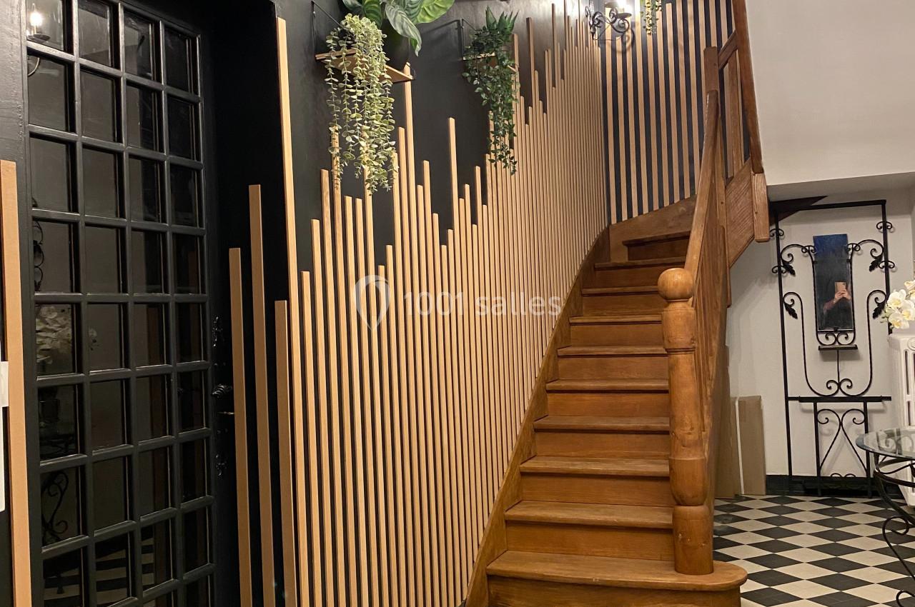 Escalier en bois avec rampe décorative, mur noir orné de plantes suspendues et sol en carrelage noir et blanc.