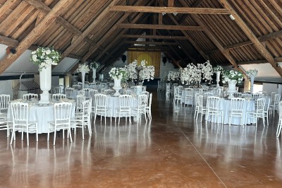 Salle de réception avec tables rondes dressées, chaises blanches et décorations florales sous une charpente en bois.