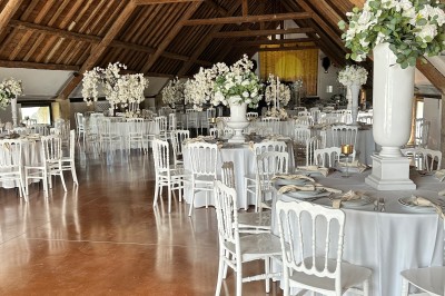 Salle de réception avec tables rondes dressées, chaises blanches et décorations florales sous une charpente en bois.