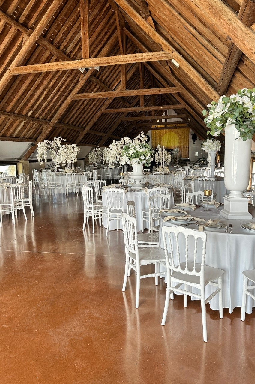 Salle de réception avec tables rondes dressées, chaises blanches et décorations florales sous une charpente en bois.