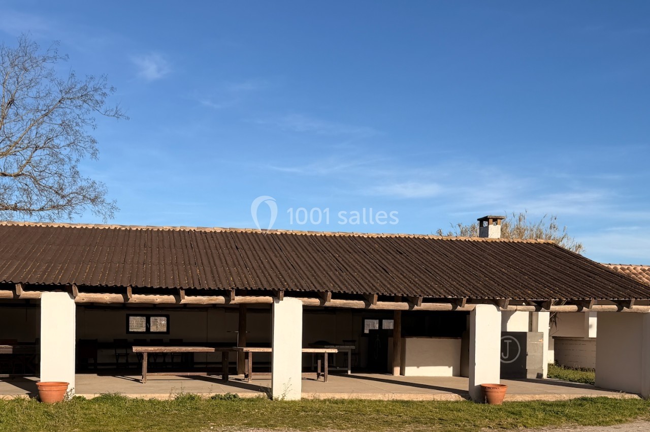 Abri extérieur avec toit en tuiles, tables et panneaux d'affichage, entouré de végétation et ciel dégagé.