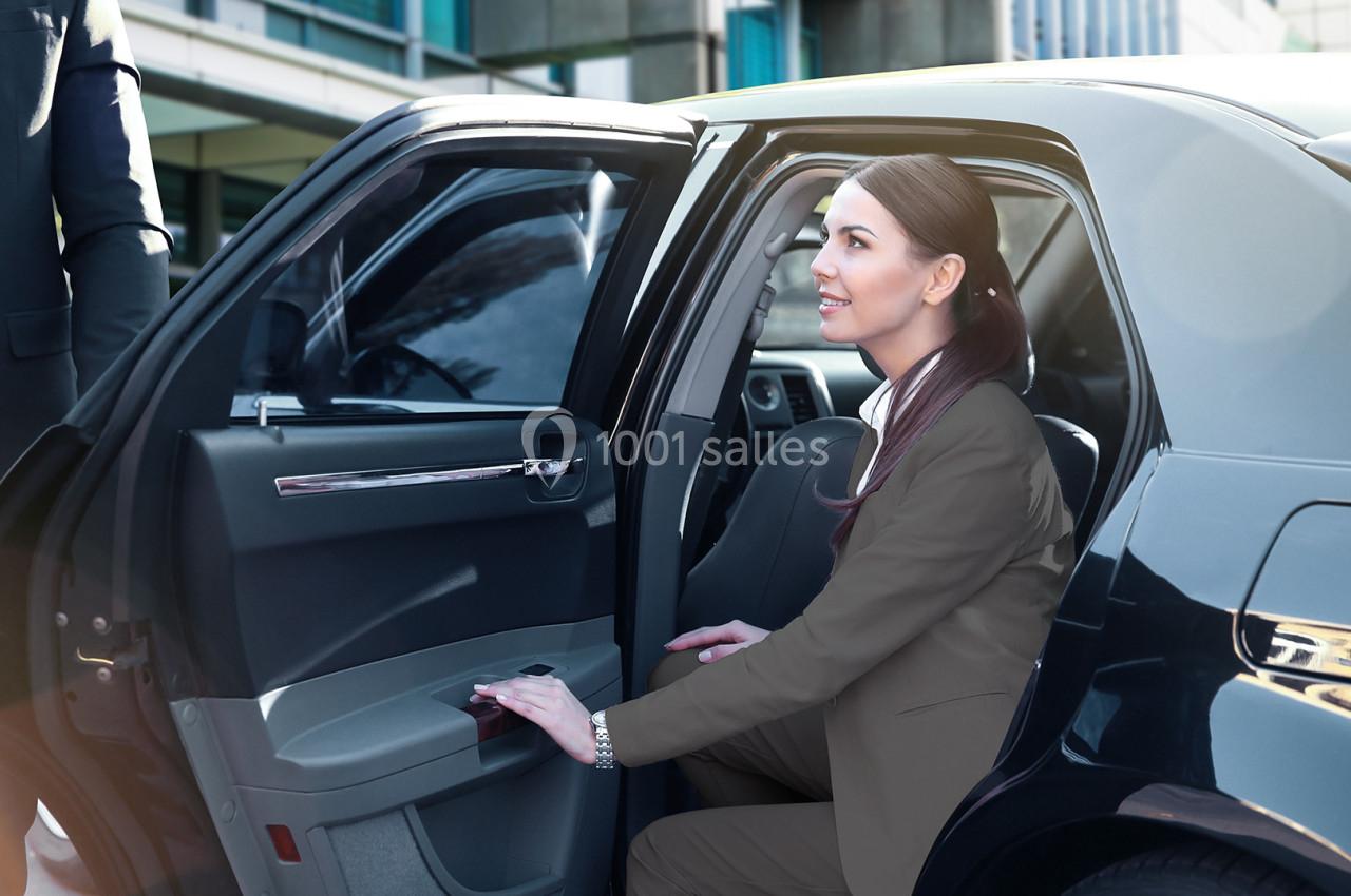 Une femme en tenue professionnelle sort d'une voiture noire, accueillie par un chauffeur en costume.