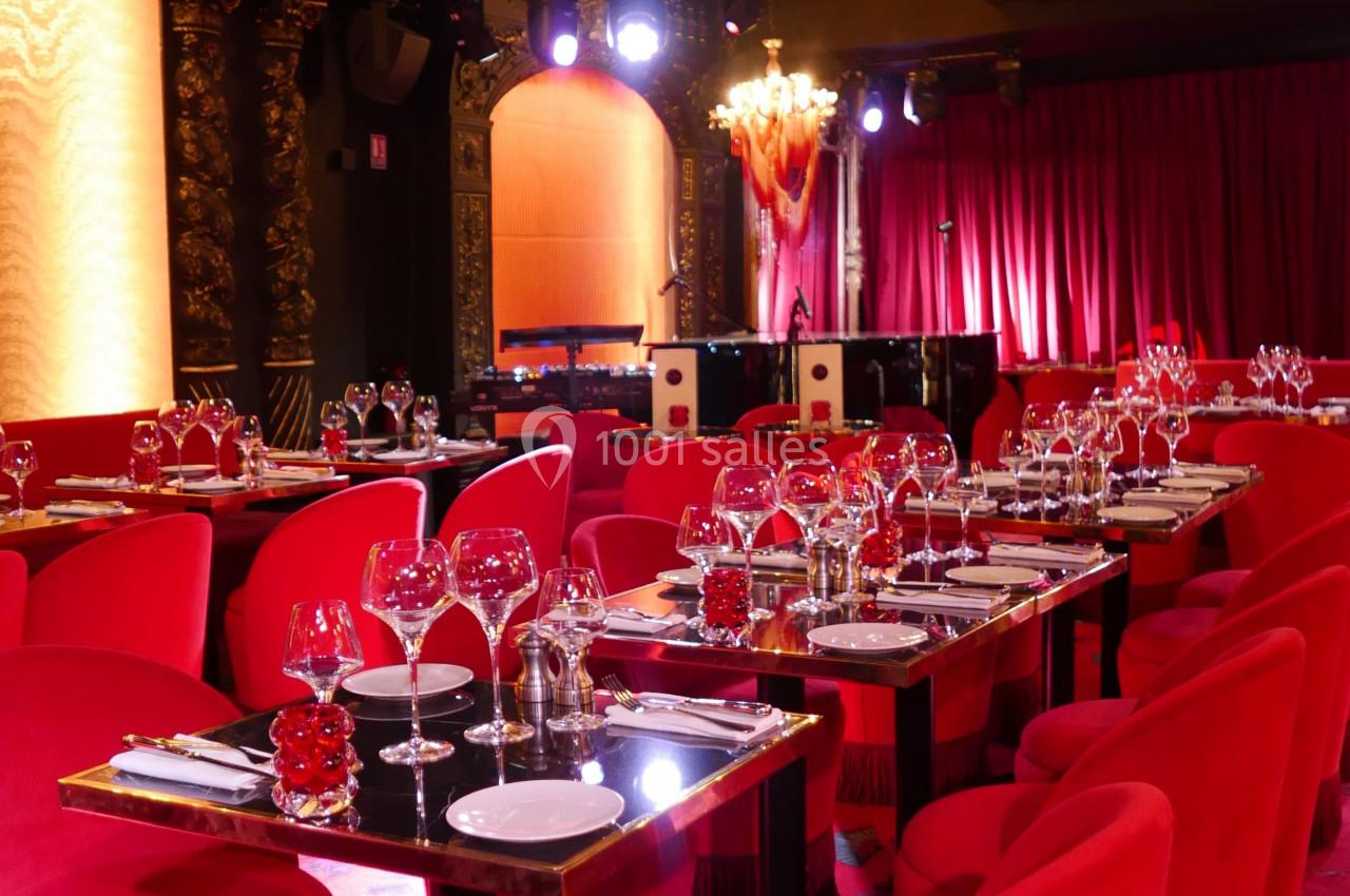 Salle de restaurant élégante avec des tables dressées, des chaises rouges et une scène éclairée en arrière-plan.