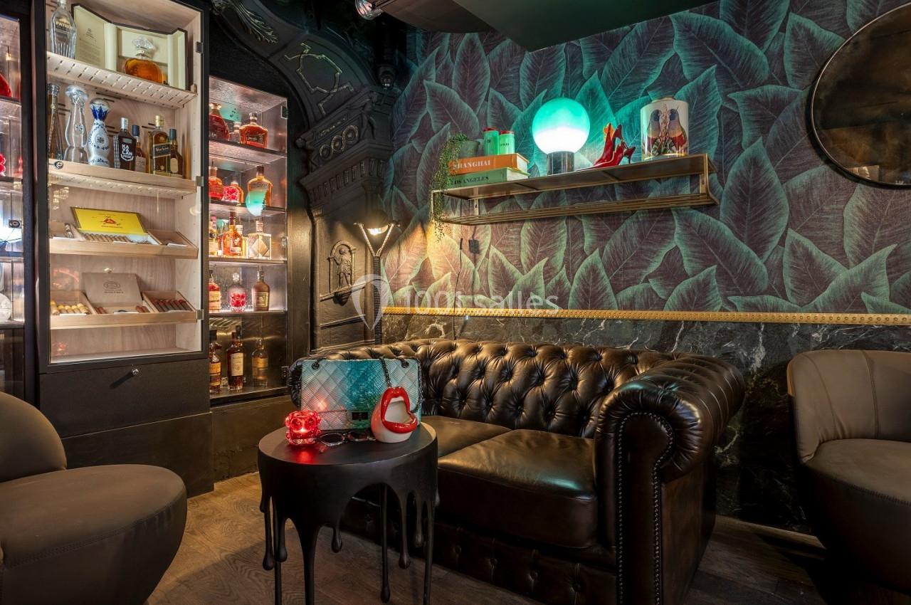 Canapé en cuir sombre dans un salon décoré de feuilles murales, avec étagères, lampe et vitrine d'alcools éclairée.