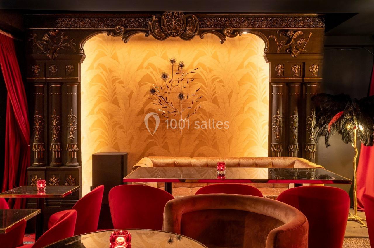 Salle élégante avec banquette beige, fauteuils rouges, décor mural doré et éclairage tamisé.