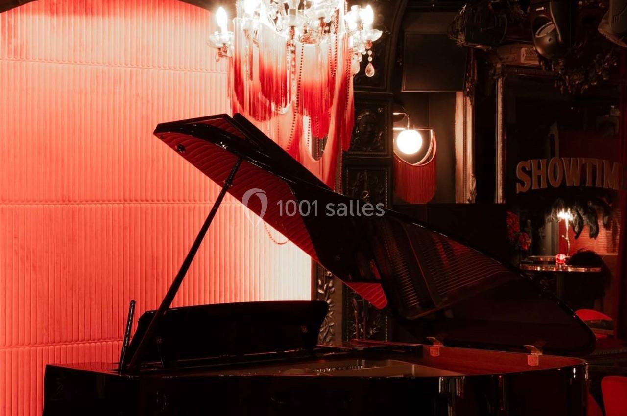Un piano à queue noir dans une salle éclairée par une lumière rouge, avec un lustre et un décor théâtral en arrière-plan.