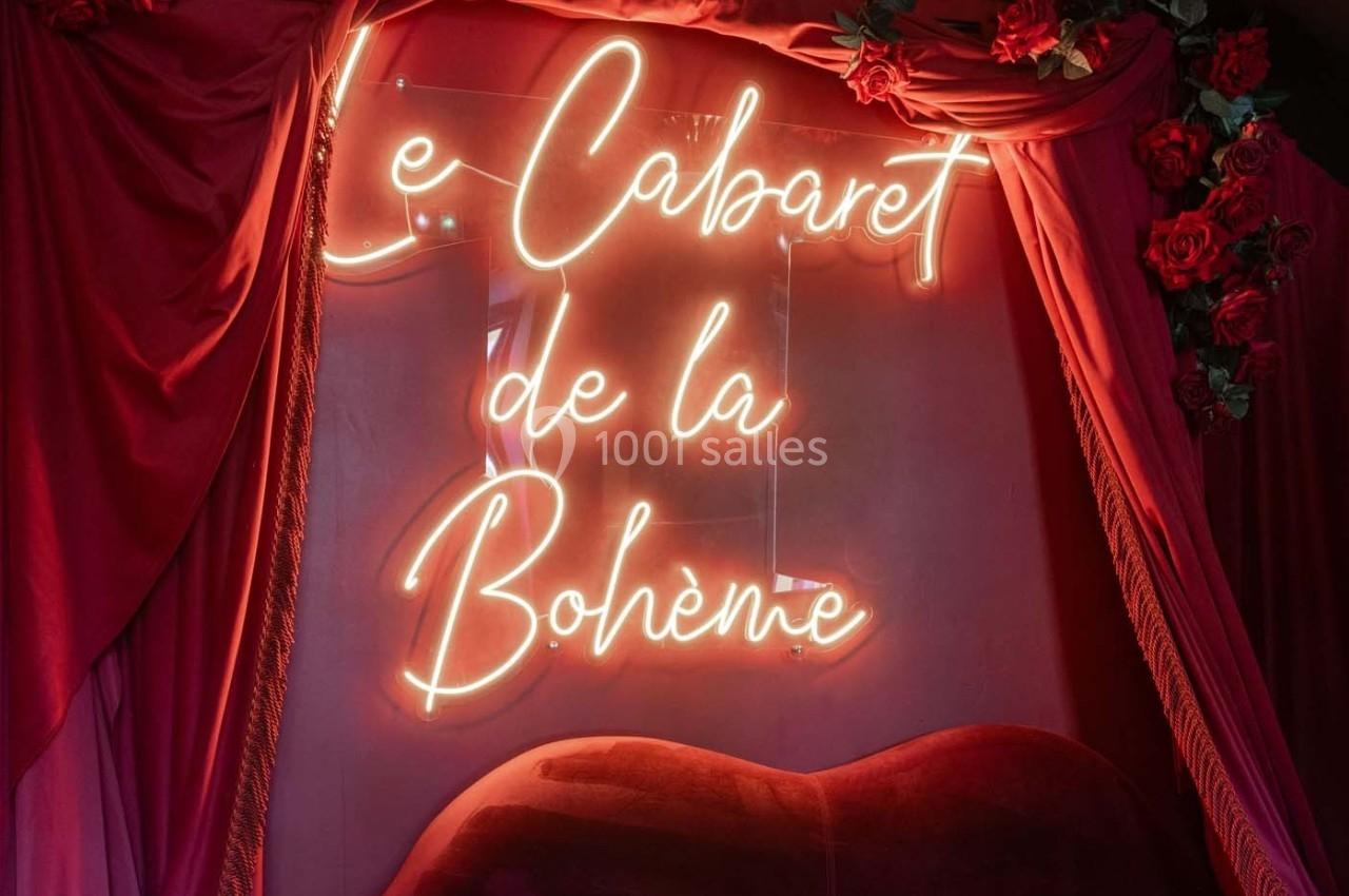 Enseigne lumineuse ’Le Cabaret de la Bohème’ entourée de rideaux rouges et de roses décoratives.