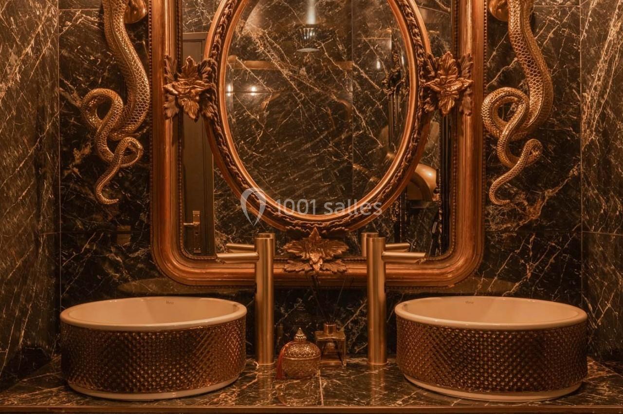 Salle de bain luxueuse avec lavabo double, miroir ovale doré et décorations serpent sur fond de marbre sombre.