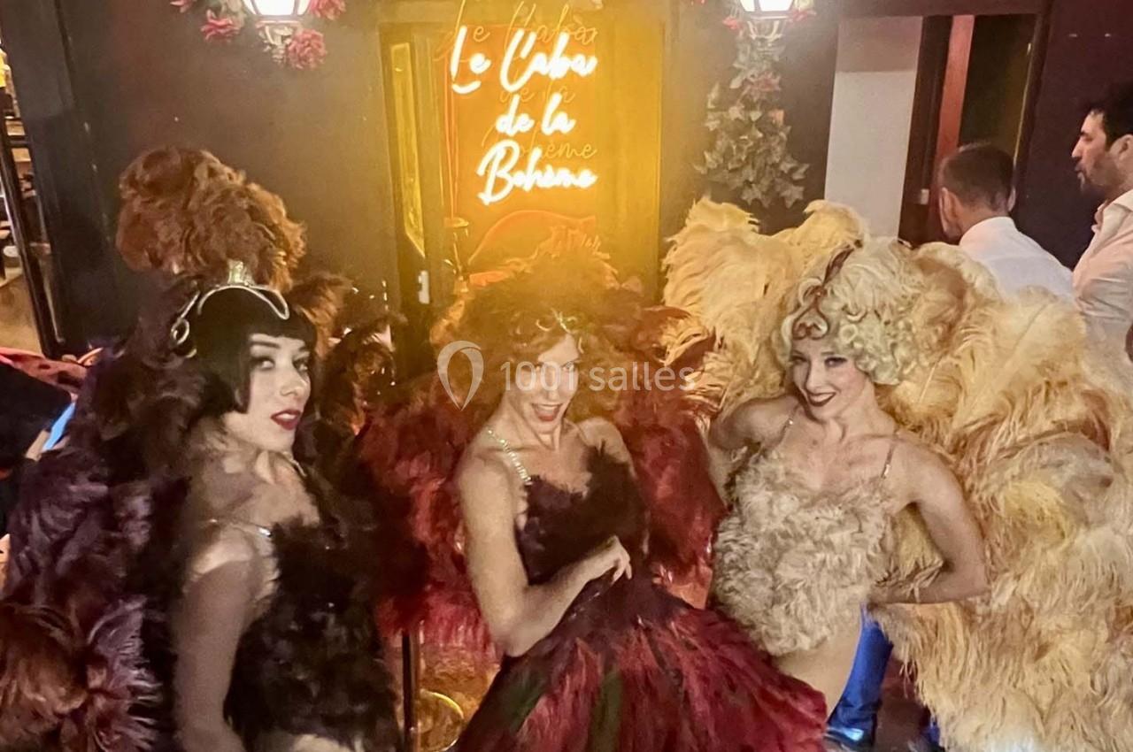 Trois danseuses en costumes à plumes colorées posent devant une entrée illuminée avec une enseigne néon.