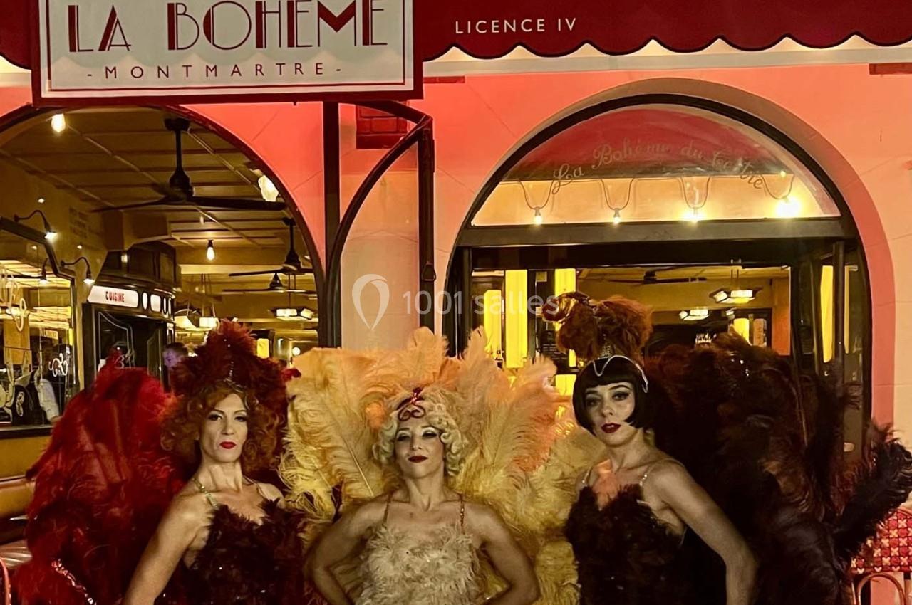 Trois danseuses en costumes à plumes colorées posent devant l'entrée éclairée du cabaret ’La Bohème’ à Montmartre.