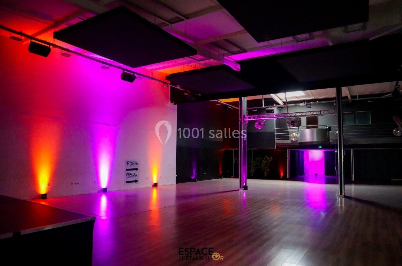 Salle spacieuse avec parquet, éclairée par des lumières colorées et équipée d'une scène et de structures métalliques.