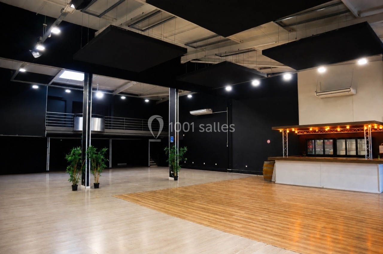 Salle spacieuse avec parquet, bar éclairé, plantes décoratives et mezzanine, éclairée par des spots au plafond.