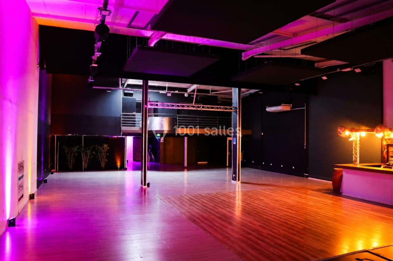 Salle spacieuse avec éclairage coloré, parquet au sol, bar à droite et mezzanine en arrière-plan.