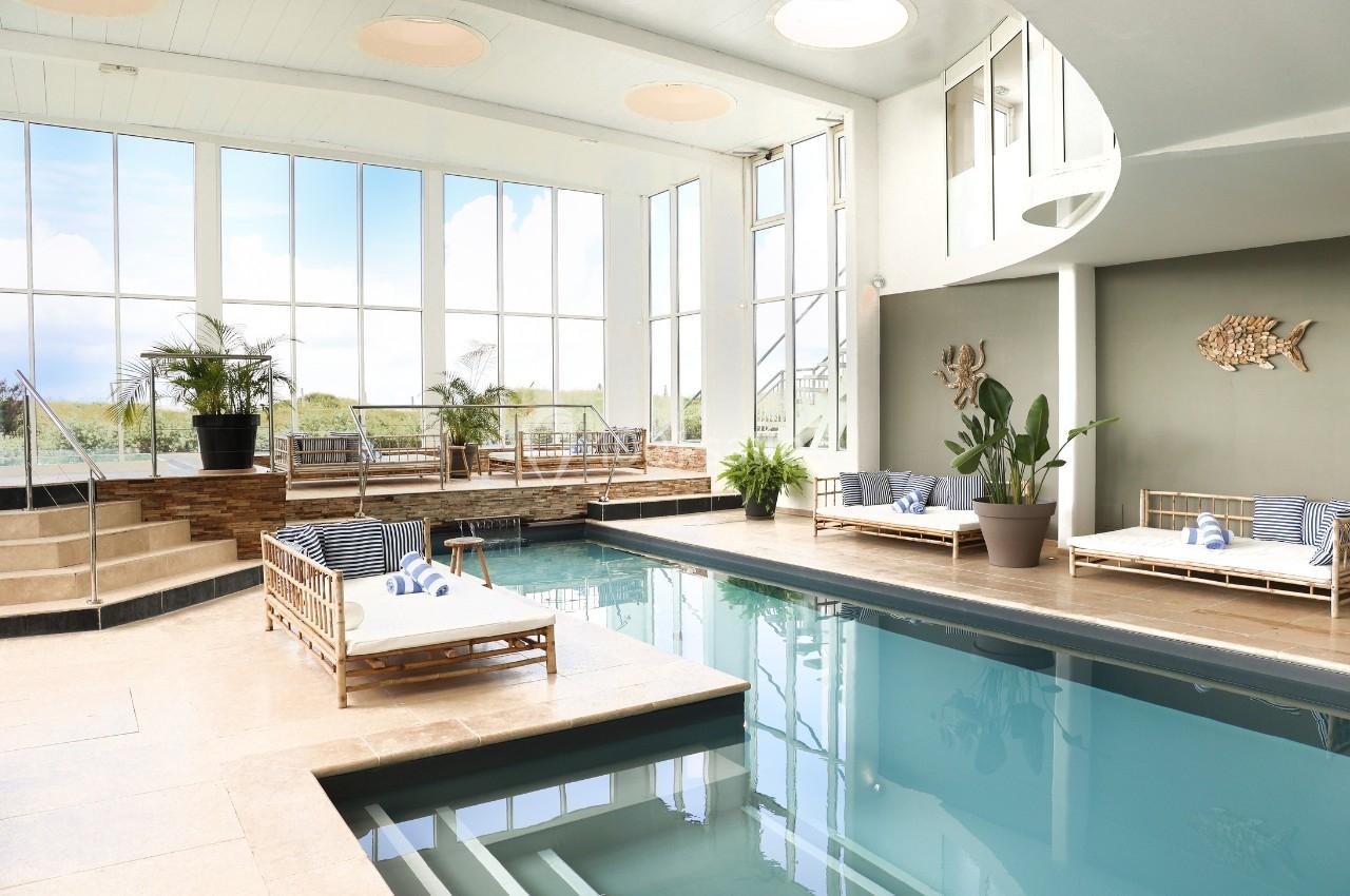 Piscine intérieure lumineuse avec transats en rotin, plantes décoratives et grandes baies vitrées donnant sur l'extérieur.