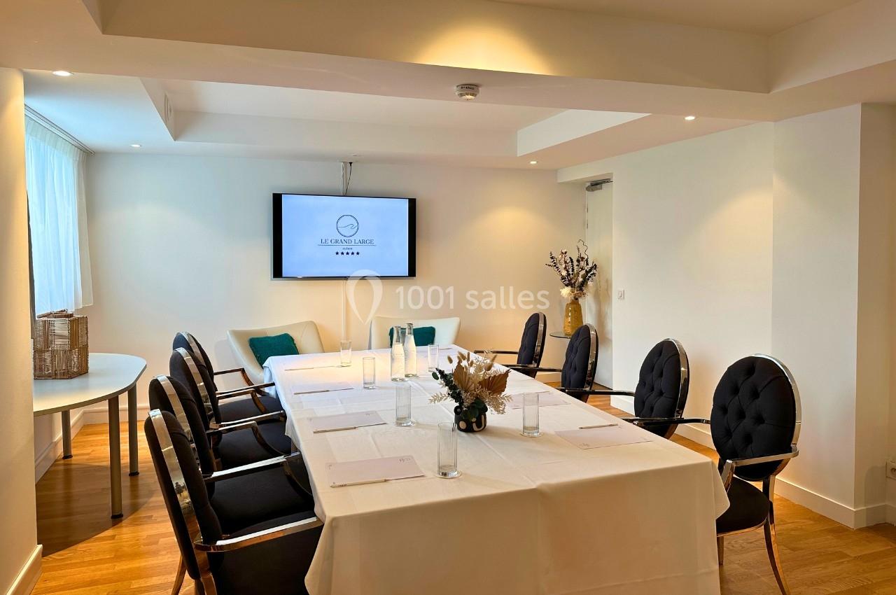 Salle de réunion lumineuse avec table rectangulaire dressée, chaises noires et écran mural allumé.
