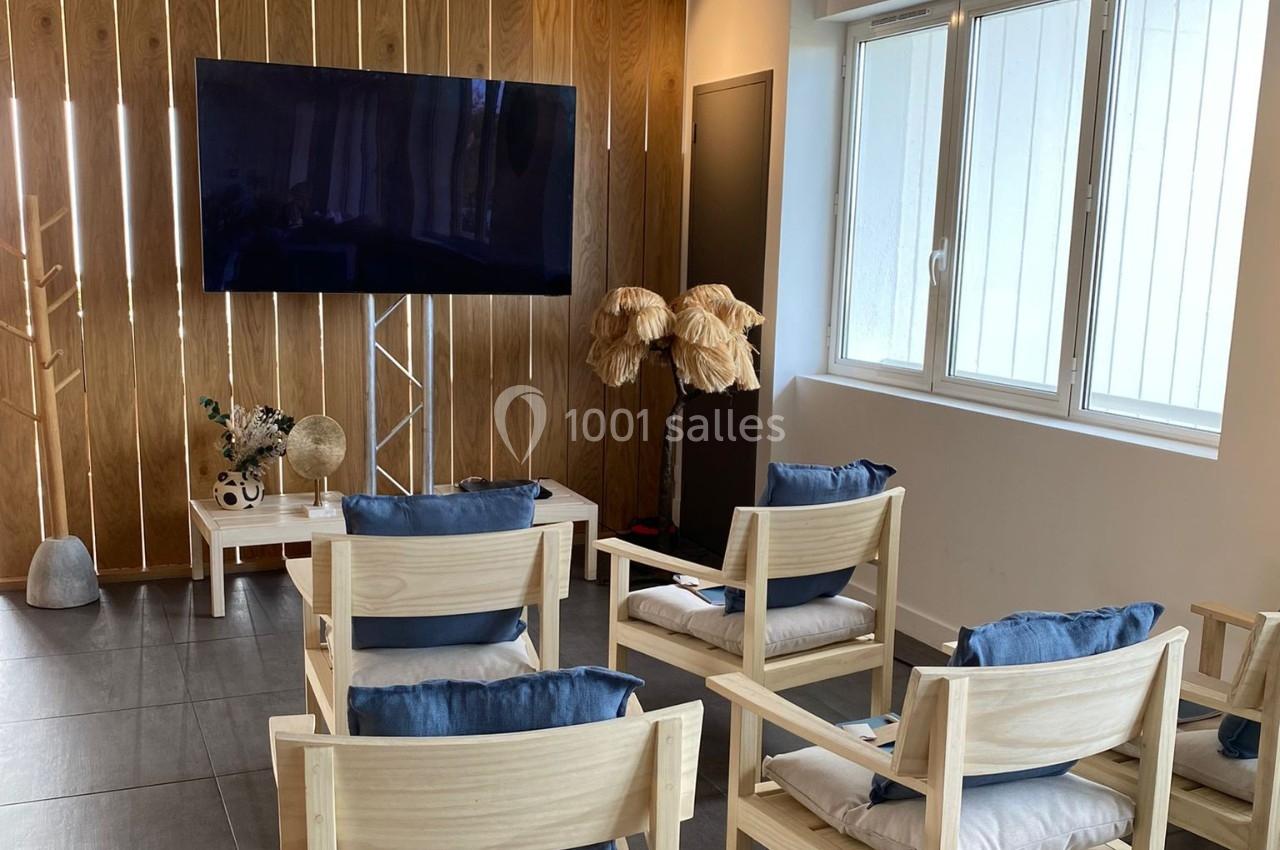 Salle lumineuse avec des chaises en bois, coussins bleus, écran mural et décoration naturelle en arrière-plan.