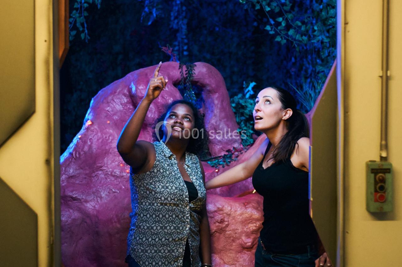 Deux femmes observent un élément lumineux dans un décor naturel avec des plantes et des rochers roses.