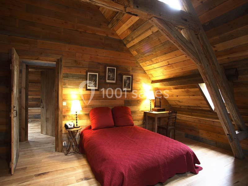 Chambre mansardée avec murs en bois, lit double avec couvre-lit rouge, bureau en bois et lumière tamisée.