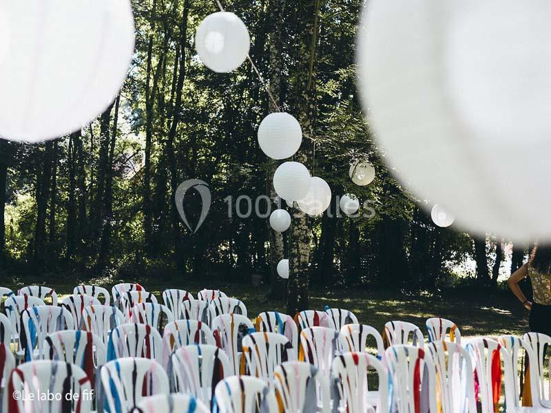 Chaises blanches décorées de rubans colorés disposées en extérieur sous des lanternes suspendues dans un cadre boisé.