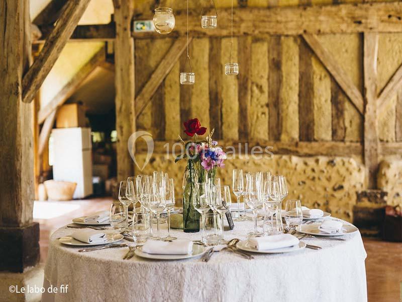 Table ronde élégamment dressée avec nappes blanches, verres, couverts et un centre de table floral dans une salle rustique.