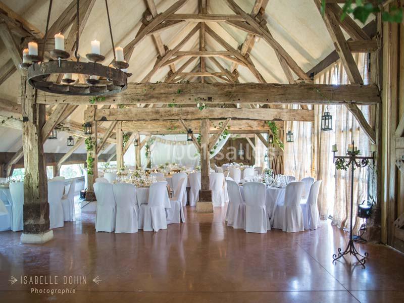 Salle de réception rustique avec poutres en bois apparentes, tables dressées et chaises blanches.