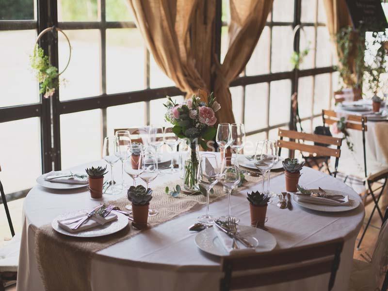 Table dressée avec nappe blanche, vaisselle élégante, bouquet de fleurs au centre et petites plantes en pot.