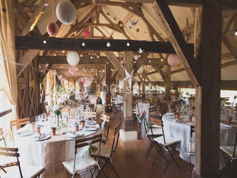 Salle de réception rustique avec poutres apparentes, tables rondes dressées et décorations suspendues.