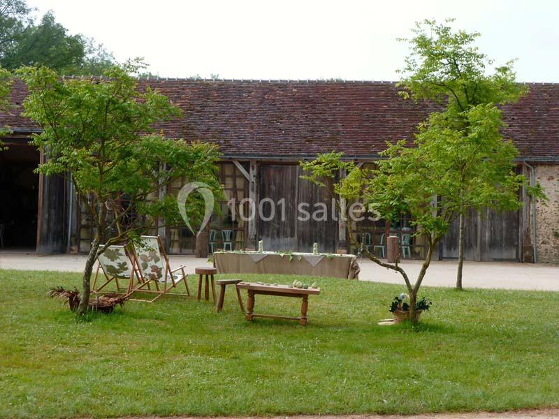 Deux arbres entourent un banc en bois et une table basse sur une pelouse devant un bâtiment en pierre et bois.