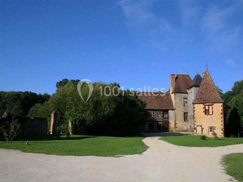 Bâtiment ancien en pierre avec toit en tuiles, entouré de verdure et d'une allée en gravier sous un ciel bleu.