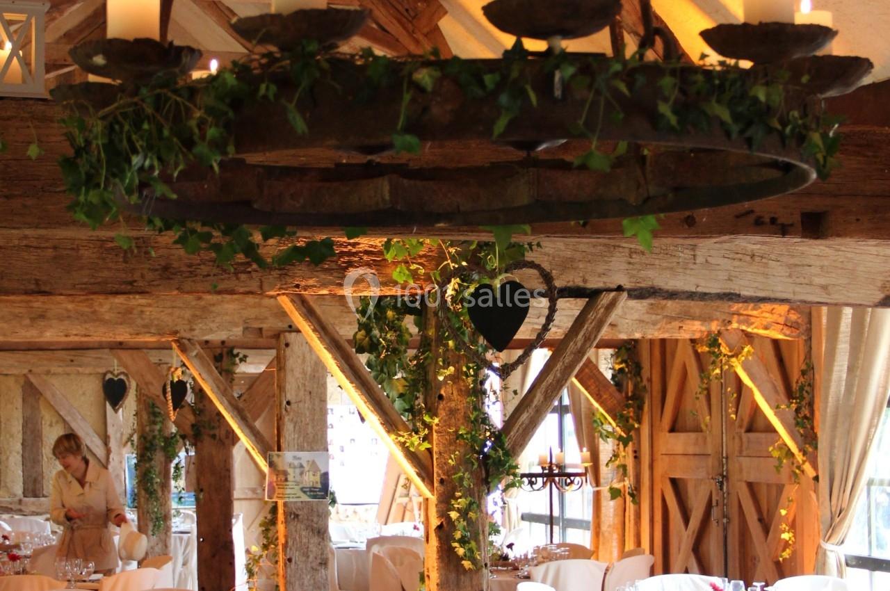 Salle de réception rustique avec poutres apparentes, tables dressées et lustre décoré de bougies et de feuillage.