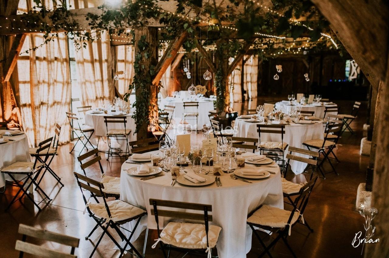 Salle de réception décorée avec des tables rondes dressées, chaises pliantes et guirlandes lumineuses suspendues.