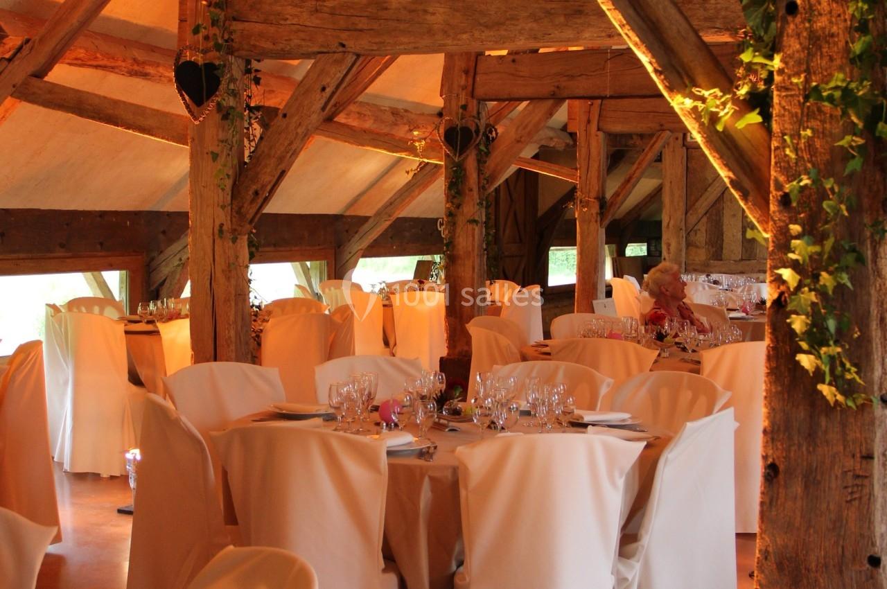 Salle de réception rustique avec poutres apparentes, tables rondes dressées et chaises couvertes de housses blanches.