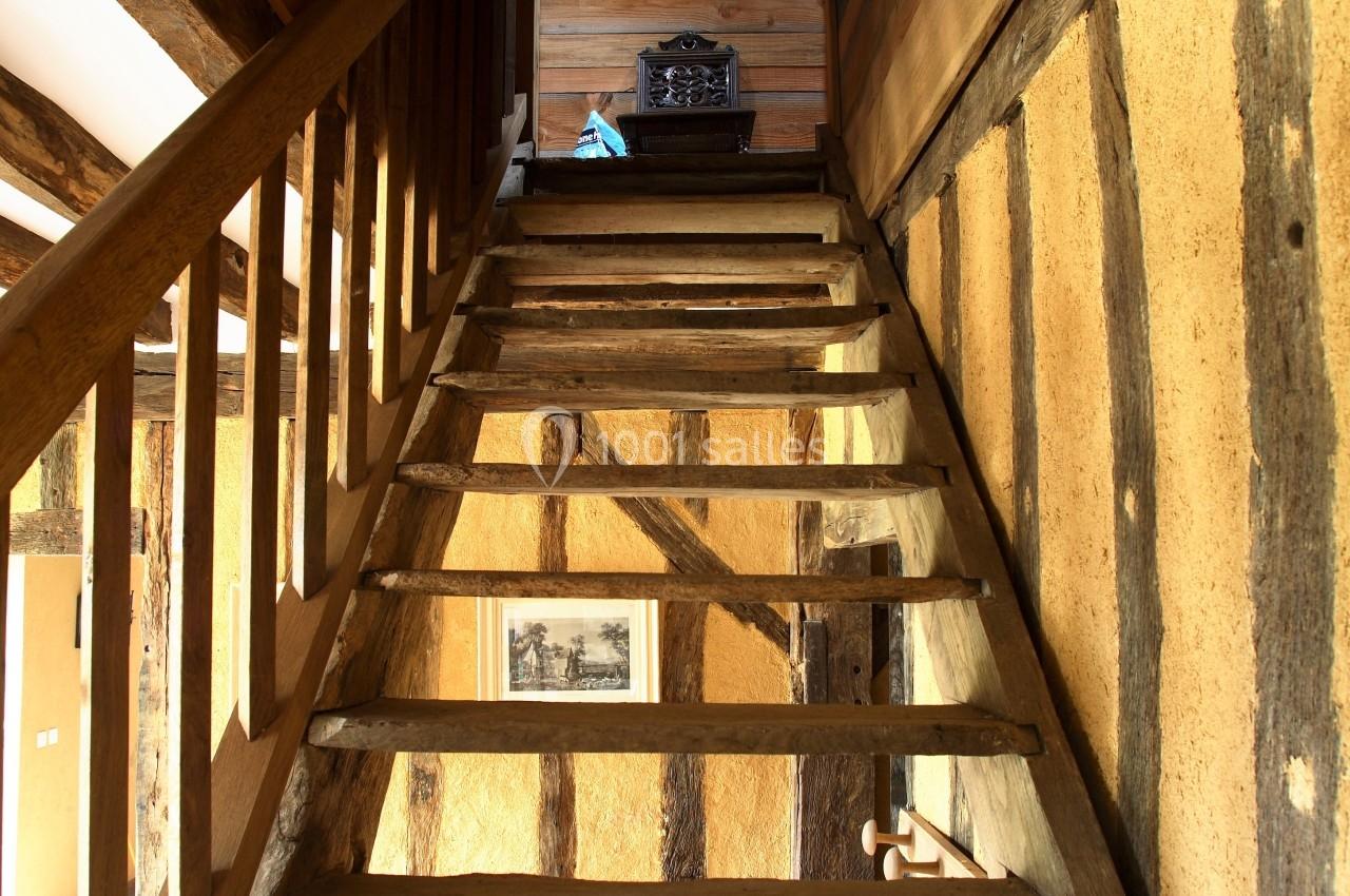 Escalier en bois rustique montant vers un étage, entouré de murs en bois et torchis dans une maison ancienne.