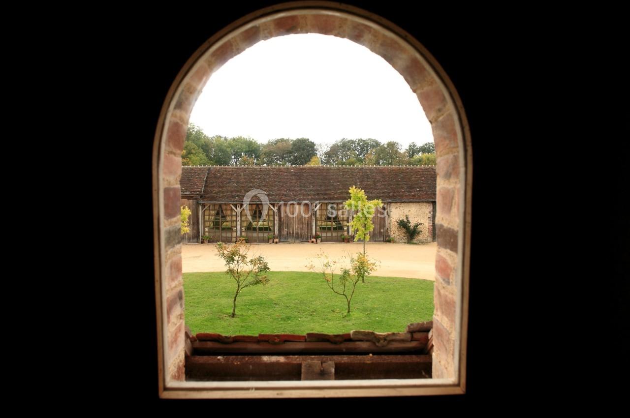 Vue d'une cour avec pelouse et bâtiments en bois, encadrée par une fenêtre en arc de briques.