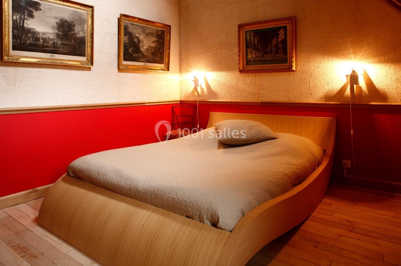 Lit double avec couverture beige dans une chambre aux murs rouges et blancs, éclairée par deux appliques murales.