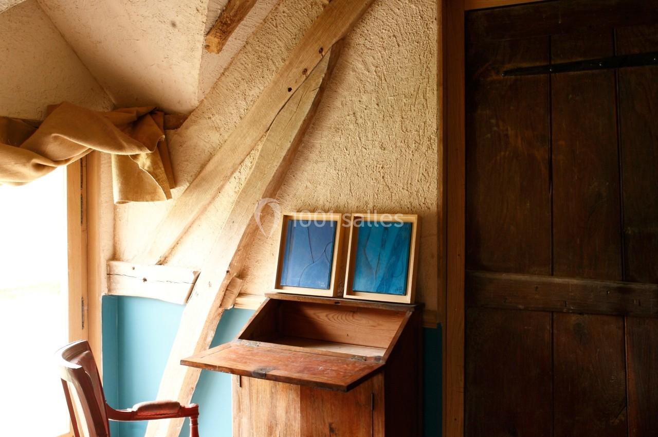 Intérieur d'une pièce rustique avec un bureau en bois, une chaise ancienne et deux cadres posés près d'une fenêtre.